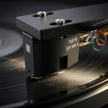 Close-up of a black Hana SL MK II vinyl record cartridge mounted on a tonearm over a spinning record, with visible red, green, and blue wire rings and white branding text on the cartridge body.