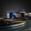 A close-up of a Hana brand blue and black cartridge on a turntable, shown resting on a spinning vinyl record against a dark studio background.