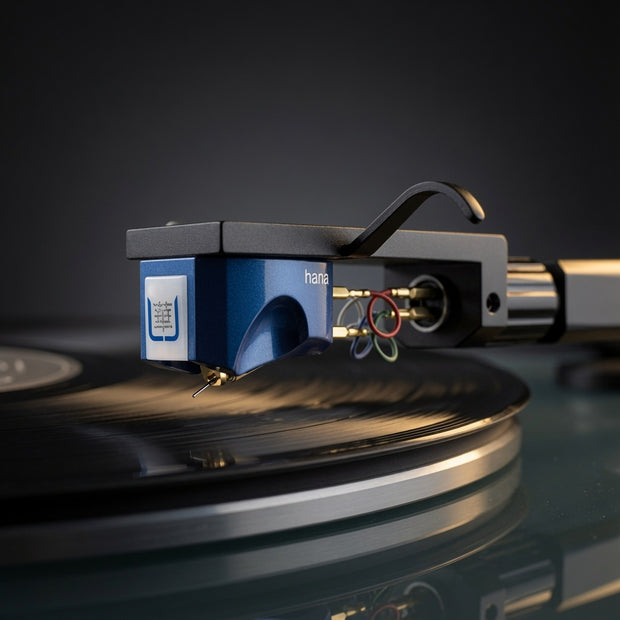A close-up of a Hana brand blue and black cartridge on a turntable, shown resting on a spinning vinyl record against a dark studio background.